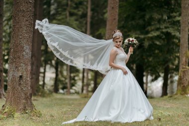 Beyaz elbiseli bir kadın saçında bir peçeyle bir ormanda duruyor. Elinde bir buket çiçek tutuyor.