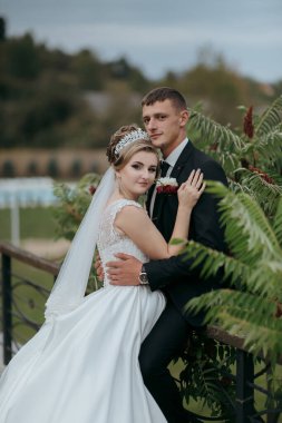 Bir gelin ve damat bir köprüde fotoğraf için poz veriyorlar. Gelin beyaz bir elbise giyiyor ve damat siyah bir takım elbise giyiyor. İkisi de gülümsüyor ve mutlu görünüyorlar. Sahne neşeli ve kutlayıcıdır.