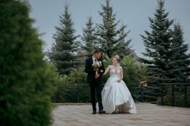 Bir gelin ve damat ormanda birlikte yürüyorlar. Gelin bir buket tutuyor ve damat elini tutuyor. Sahne romantik ve mutlu.