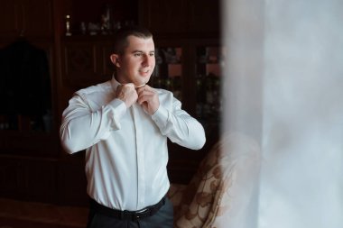 Beyaz gömlekli bir adam pencerenin önünde giyiniyor. Sahne rahat ve rahattır. Adam gün boyu giyinir.