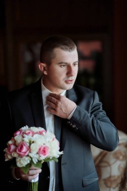 Takım elbiseli bir adam elinde bir buket çiçek tutarken kravatını düzeltiyor. Zarafet ve entelektüellik anlayışı, çünkü adam resmi giyinmiş.