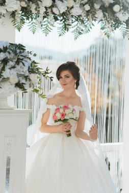 Bir gelin elinde bir buket çiçekle beyaz bir kemer yolunun önünde duruyor.