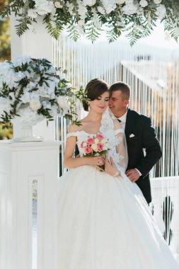 Gelin ve damat, beyaz bir kemerin altında ellerinde bir buket çiçekle duruyorlar. Sahne büyük ihtimalle bir düğün merasimi, ve çift bir resim için poz veriyor.