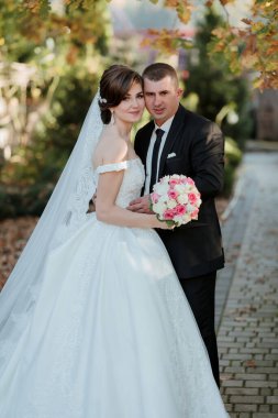 Bir gelin ve damat bir buket çiçekli fotoğraf için poz veriyorlar. Gelin beyaz bir elbise giyiyor ve damat siyah bir takım elbise giyiyor. Sahne mutlu ve kutlama dolu.