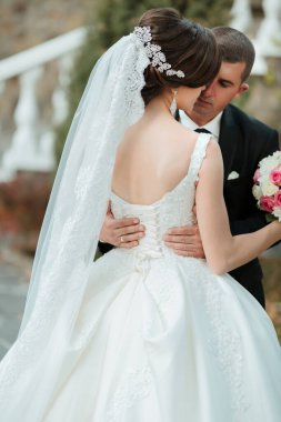 Gelin ve damat birlikte duruyorlar. Gelin duvağı olan beyaz bir elbise ve damat siyah bir takım elbise giyiyor.