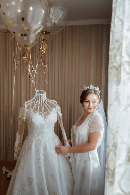 Kafasında taç olan beyaz bir elbisenin önünde duran bir kadın. Elinde elbise askısı ve balon var. Sahne şenlikli ve kutlamalı, büyük ihtimalle düğün ya da diğer özel durumlar için.