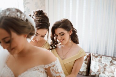 Üç kadın düğün için hazırlanıyor. İçlerinden biri sarı bir elbise giyiyor. Diğer iki kadın giyinmesine yardım ediyor.