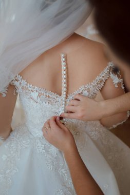 Bir kadın düğüne hazırlanıyor ve elbisesinin düğmelerini ayarlıyor. Elbise beyaz ve dantel tasarımı var. Kadına başka biri yardım ediyor. Elbisesini yukarı kaldırıyor.