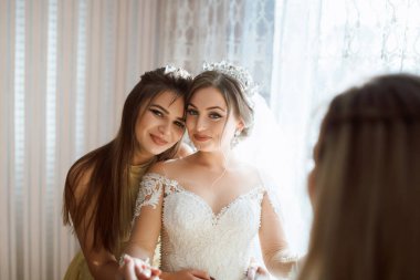İki kadın fotoğraf için poz veriyor, biri gelinlik giyiyor. Diğer kadın gelinin elini tutuyor.