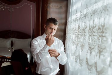 Beyaz gömlekli bir adam giyinmeye hazırlanıyor. Penceredeki yansımasına bakıyor.