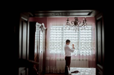 Bir adam pembe duvarlı bir odada giyiniyor. Beyaz gömlek ve kırmızı pantolon giyiyor.