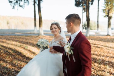 Bir gelin ve damat parkta birlikte yürüyorlar. Gelin bir buket çiçek tutuyor ve damat kırmızı bir elbise giyiyor. Sahne mutlu ve romantik.