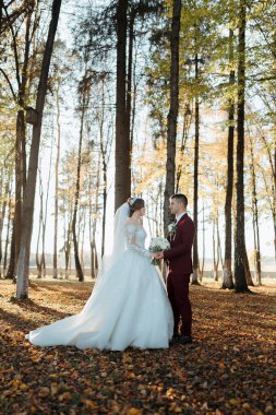 Bir gelin ve damat ağaçlar ve yapraklarla çevrili bir ormanda duruyor. Gelin beyaz bir elbise giyiyor ve damat kırmızı bir takım elbise giyiyor. El ele tutuşup gülümsüyorlar, mutlu görünüyorlar.