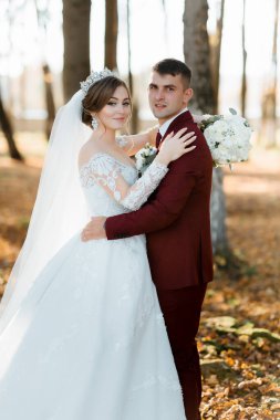 Bir gelin ve damat ormanda fotoğraf çektirmek için poz veriyorlar. Damat kırmızı bir takım giyerken gelin beyaz bir elbise ve taç giyiyor. El ele tutuşup gülümsüyorlar.