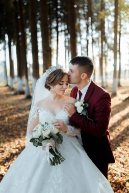 Bir gelin ve damat ormanda sarılıyor. Gelin beyaz bir elbise giyiyor ve damat kırmızı bir takım elbise giyiyor.