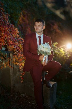 Kırmızı takım elbiseli ve beyaz kravatlı bir adam elinde bir buket çiçek tutuyor. Adam taş bir çıkıntının üzerinde duruyor ve çiçekler bembeyaz. İmajın romantik ve zarif bir havası var.