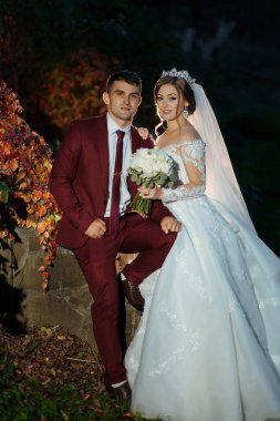 Bir gelin ve damat duvarın önünde fotoğraf için poz veriyorlar. Gelin beyaz bir elbise giyiyor ve damat kırmızı bir takım elbise giyiyor.