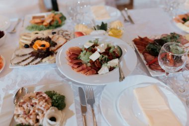 İçinde salata, et ve peynir de dahil çeşitli yiyecek ve içeceklerin olduğu bir masa. Masa çatal bıçak ve bardaklarla dolu. Masada birkaç şişe ve bardak var.