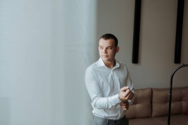 Beyaz gömlekli bir adam bir pencerenin önünde duruyor, sokağa bakıyor. Gri bir takım elbise ve gümüş bir bilezik takıyor. Sahne sakin ve derin derin.