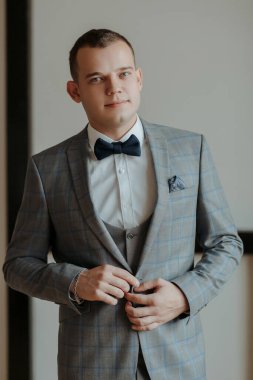 Takım elbiseli ve kravatlı bir adam mavi bir papyon takıyor. Beyaz bir duvarın önünde duruyor.