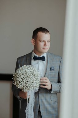 Elinde bir buket çiçek tutan takım elbiseli bir adam. Adam mavi bir papyon ve gri bir ceket giyiyor.