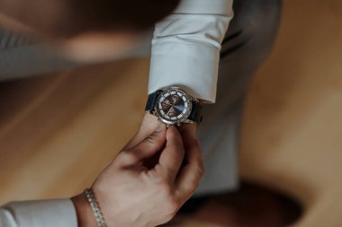 Bir adam saat takıyor ve ayarlıyor. Saat gümüş ve siyah. Adam beyaz gömlek ve gri pantolon giyiyor.