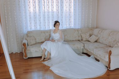 Beyaz elbiseli bir kadın oturma odasındaki bir kanepede oturur. Kanepe beyaz yastıklarla kaplı ve duvarında bir ayna var. Oda aydınlık ve hoş, davetkar bir atmosferi var.