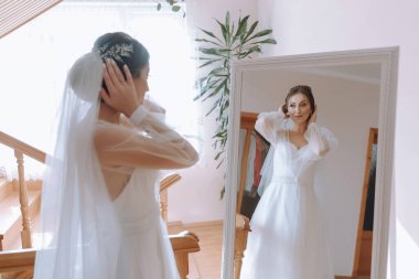 Bir kadın düğünü için hazırlanıyor ve aynada kendine bakıyor. Beyaz bir elbise ve duvak takıyor.