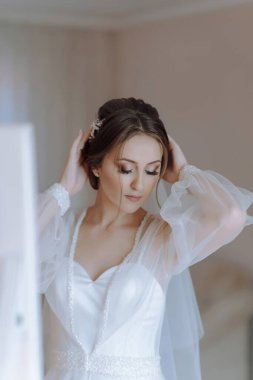 Bir kadın düğün için hazırlanıyor, gelinliğini ve duvağını giyiyor. Aynanın önünde duruyor, saçını ve makyajını ayarlıyor.