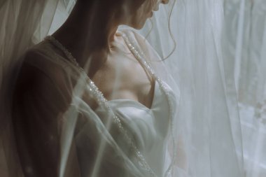 Beyaz elbiseli bir kadın perdenin önünde duruyor. Elbise beyaz ve dantel bir yakası var. Zarafet ve resmiyet kavramı