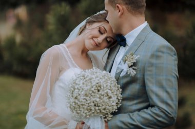 Bir gelin ve damat sarılıyor ve gelin bir buket tutuyor. Sahne mutlu ve romantik.