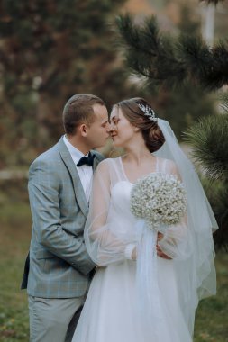 Bir gelin ve damat ormanda öpüşüyor. Gelin elinde bir buket çiçek tutuyor.