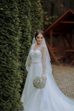 Beyaz elbiseli bir kadın çalılığın önünde duruyor. Elinde bir buket çiçek tutuyor.