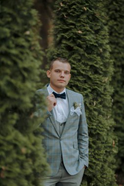 Takım elbiseli ve kravatlı bir adam çalıların önünde duruyor. O, mavi bir takım ve beyaz bir gömlek giyiyor.