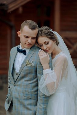Bir gelin ve damat ahşap bir binanın önünde fotoğraf için poz veriyorlar. Gelin beyaz bir elbise giyiyor ve damat bir takım elbise giyiyor. İkisi de gülümsüyor ve birbirlerine sarılıyorlar.