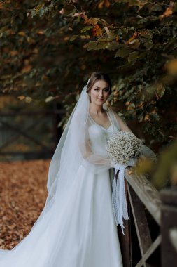 Bir gelin elinde bir buket çiçekle çitin önünde duruyor. Beyaz bir elbise ve duvak takıyor.