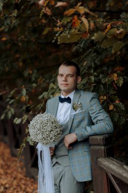 Takım elbiseli ve kravatlı bir adam elinde bir buket çiçekle köprüde duruyor. Mavi bir ceket ve papyon takıyor.