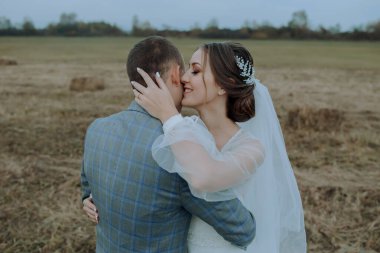 Bir gelin ve damat tarlada sarılıyor. Gelin beyaz bir peçe takıyor ve damat mavi bir takım giyiyor.