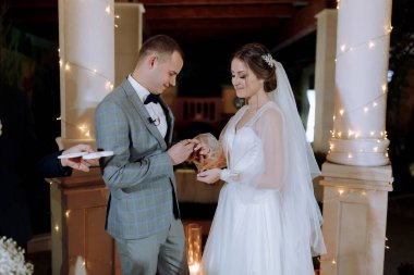 Bir erkek ve bir kadın evleniyor ve erkek kadına bir yüzük vermek üzere. Gelin beyaz bir elbise giyiyor ve damat mavi bir takım elbise giyiyor. Ortam romantik ve mutlu.