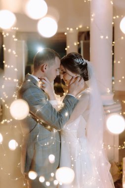 Bir gelin ve damat ışıklandırılmış bir arka planda kucaklaşıyorlar. Gelin beyaz bir elbise giyiyor ve damat bir takım elbise giyiyor. Sahne romantik ve samimi, aşkı yakalıyor.