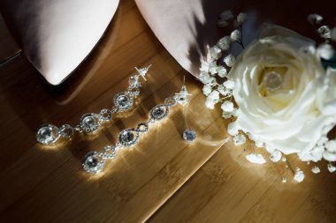 Tahta bir masanın üzerinde bir çift küpe ve bir gül var. Küpeler gümüş ve elmas desenli. Gül beyazdır ve küçük bir beyaz çiçek kümesine sahiptir. Sahne zarif ve romantik.