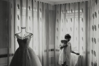 Bir kadın düğün için hazırlanıyor ve arka planda bir elbise asılı duruyor. Oda loş bir şekilde aydınlanıyor ve perdeler çekiliyor, mahremiyet ve yakınlık hissi yaratıyor.