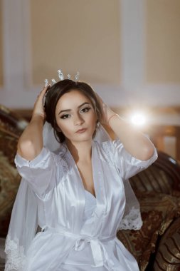 Beyaz cüppeli bir kadın kanepede oturuyor ve tacını ayarlıyor. Şık ve sofistike bir kadın düğün kıyafeti giymiş ve taç takıyor.