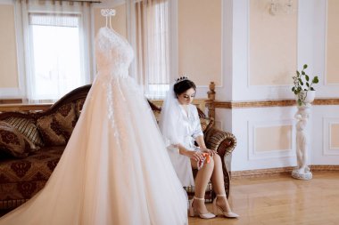 Bir gelin, bir kanepede gelinliğin önünde oturuyor. Elbise beyaz ve uzun bir treni var. Gelin elinde bir buket tutuyordu. Sahne, kanepeli, sandalyeli bir odada geçiyor.