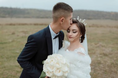 Bir erkek beyaz gelinlik giyerken bir kadının yanağını öper. Gelin elinde bir buket beyaz gül tutuyor.