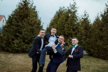 Takım elbiseli dört adam bir resim için poz verirken, bir adam diğerini kaldırıyor. Sahne neşeli ve eğlenceli, çünkü erkekler birbirlerinin arkadaşlığından zevk alıyor ve iyi vakit geçiriyorlar.