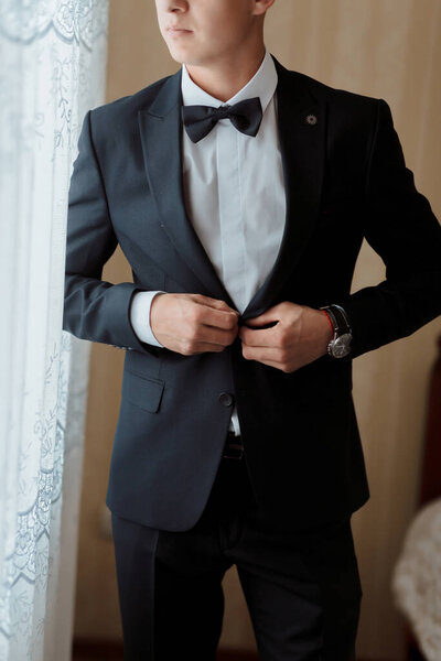 A man in a suit is getting ready to go out. He is wearing a black suit and a white shirt. He is adjusting his tie and looking out the window. Scene is formal and serious