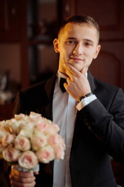 Takım elbiseli ve kravatlı bir adam elinde bir buket çiçek tutuyor. Yüzünde bir gülümsemeyle kameraya bakıyor.