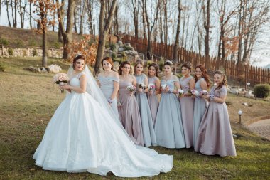 Bir gelin ve nedimeleri tarlada fotoğraf çektirmek için poz veriyorlar. Gelin beyaz bir elbise giyiyor ve elinde bir buket çiçek tutuyor.