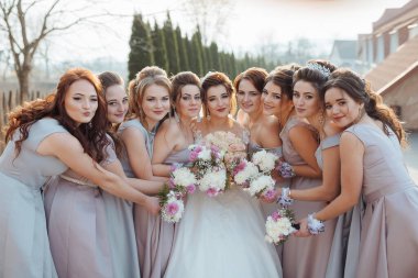 Bir grup kadın bir resim için poz veriyor, bir tanesi elinde bir buket çiçek tutuyor. Sahne neşeli ve kutlama dolu, çünkü kadınlar düğün kıyafetleri giymişler ve grup fotoğrafı için poz veriyorlar.